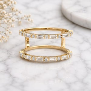 May include: A double-band gold ring featuring two parallel bands connected by vertical gold bars. Each band is set with alternating baguette-cut and round brilliant-cut clear stones arranged in a continuous pattern around the entire circumference.