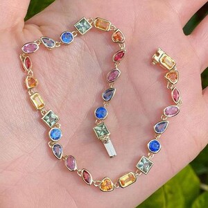 May include: A gold chain bracelet featuring a variety of multi-coloured gemstones in diverse shapes including round, square, pear, and marquise cuts. The bracelet includes a secure box clasp closure and displays stones in shades of blue, pink, red, and yellow.