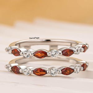 Peut inclure: Deux bagues en argent ornées de pierres précieuses orange en forme de marquise et de pierres blanches rondes alternées. Les bagues sont présentées sur une surface blanche avec un arrière-plan flou.