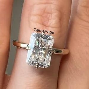 Può includere: Un anello con fascia in oro e una grande gemma trasparente di forma rettangolare. La gemma è incastonata in una montatura a quattro griffe. L'anello è su un dito. Il testo "GemsPage" è visibile.