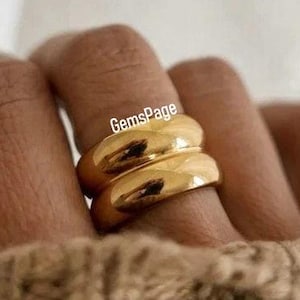 May include: Two gold-colored rings stacked on a finger. The rings have a smooth, rounded design. The image is set against a neutral background, with the rings and the hand in focus. The rings are on a hand wearing a tan sweater. The text "GemsPage" is visible.