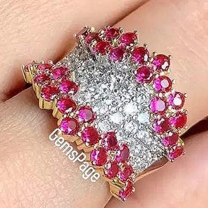 Op de afbeelding: Een goudkleurige ring met een cluster van kleine, ronde, rode edelstenen rond een centraal gebied van kleinere, heldere edelstenen. De ring heeft een uniek, getextureerd ontwerp.