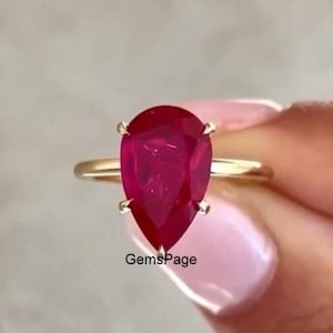 Peut inclure: Une bague en or avec une pierre précieuse rouge foncé en forme de poire montée sur un sertissage à quatre griffes. L'anneau est fin et en or poli. Le texte GemsPage est imprimé en petites lettres noires sous la pierre sur l'anneau.