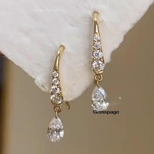 Könnte beinhalten: Ein Paar goldene Ohrhänger mit birnenförmigen, klaren Edelsteinen und kleineren, klaren Edelsteinen entlang der Ohrstecker. Die Ohrringe liegen auf einer weißen Oberfläche. Gemspace.
