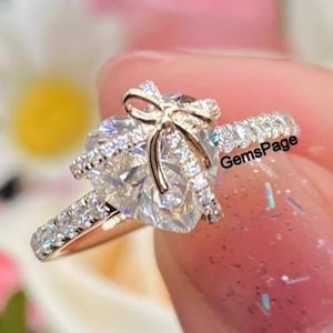 May include: A gold engagement ring featuring a pear-cut diamond and a bow design. The band is set with smaller diamonds. The ring is held up against a blurred floral background. The text "GemsPage" is visible.