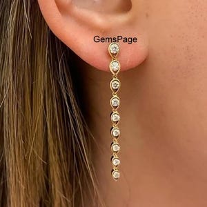以下が含まれることがあります： 小さな丸い透明な宝石が連なったゴールドのイヤリング。イヤリングは耳からぶら下がっています。耳には「GemsPage」の文字が見えます。