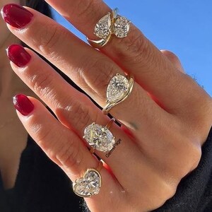May include: Four gold rings featuring clear faceted stones in various shapes including pear, oval, and heart cuts. The rings are worn on four fingers, showcasing a mix of solitaire and multi-stone designs with polished yellow gold bands.