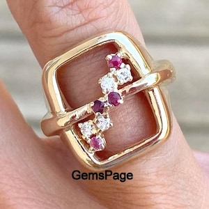 May include: A gold-toned ring with a square design, featuring a cluster of small red and white gemstones. The ring is worn on a finger, showcasing its intricate details and craftsmanship. The text "GemsPage" is visible.