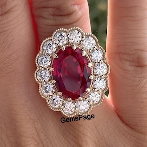 May include: A gold ring with a large, oval ruby at its centre, encircled by a halo of smaller, clear gemstones. The ring has a scalloped edge. The text "GemsPage" is visible on the ring.