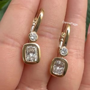 May include: A pair of gold-toned earrings with a rectangular, clear gemstone at the bottom and a smaller, round gemstone above. The earrings have a lever back closure. The text "GemsPage" is visible.