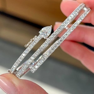 Puede incluir: Un brazalete de diamantes con un diamante en forma de pera y múltiples filas de diamantes más pequeños. El brazalete es sostenido por una persona. El texto "GemsPage" es visible.