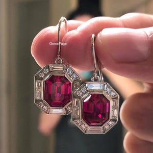 Pode incluir: Um par de brincos pendentes de tom prateado com uma grande pedra central octogonal rosa-escura rodeada por um halo de pequenas pedras de destaque claras, retangulares e redondas. Os brincos têm fecho de gancho e o texto GemsPage é visível.