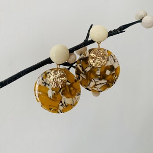 Può includere: Un paio di orecchini rotondi con motivo dorato e bianco con un accento dorato. Gli orecchini sono rotondi e hanno una perla bianca nella parte superiore.