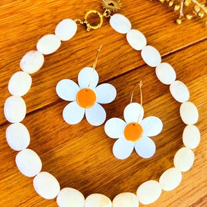 Peut inclure: Un collier de perles blanches ovales avec un fermoir doré et des boucles d'oreilles assorties en forme de marguerite. Les boucles d'oreilles ont des pétales blancs et des centres orange. L'ensemble est présenté sur une surface en bois.