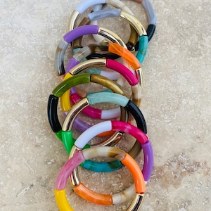 Peut inclure: Une pile de bracelets colorés avec des accents dorés. Les bracelets sont faits de différents matériaux, dont le plastique, le bois et le métal. Les bracelets sont disposés en pile, le plus grand bracelet en bas et le plus petit en haut.