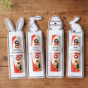 Könnte beinhalten: Vier Oster-Geschenkanhänger mit einer rot-weißen Süßwarenriegel. Jeder Anhänger hat ein anderes Design: Hasenohren, ein Hase, ein Ei und ein Hase. Jeder Anhänger hat ein weißes Band mit deutschem Text.