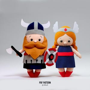May include: Two felt dolls depicting a Viking and a Valkyrie. The Viking doll has a large orange beard, a horned helmet, and holds an axe. The Valkyrie doll has red hair, a winged helmet, and a shield. The dolls are made of felt and have a whimsical design.