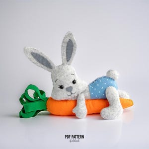 Könnte beinhalten: Ein handgefertigtes Häschen-Spielzeug aus Filz mit grau-weißem Fell, das blaue Shorts mit Punkten trägt und auf einer orangefarbenen Karotte liegt. Die Karotte hat ein grünes Band. Der Text "PDF PATTERN by belarba" steht unten.