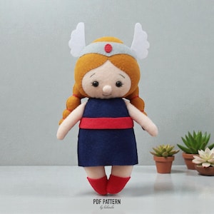 May include: A handmade felt doll with a light skin tone, wearing a navy blue dress with a red belt and red boots. The doll has orange hair in braids, a silver headband with a red gem, and white wing accents. The text "PDF PATTERN by belarinda" is at the bottom.