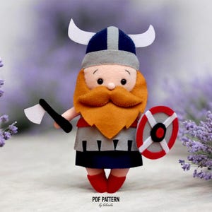 May include: A handmade felt Viking doll with a navy blue helmet, gray accents, and white horns. The doll has an orange beard, a red and white shield, and an axe. The doll is wearing a gray and blue tunic and red boots. The text "PDF PATTERN by lilaroba" is at the bottom.