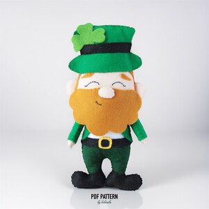 May include: A felt leprechaun doll with a green hat adorned with a shamrock, a ginger beard, and a green outfit. The doll has a smiling face and black shoes. The text "PDF PATTERN by belaruba" is at the bottom.