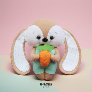 May include: A handmade felt bunny doll holding a carrot. The bunny has brown and white fur, large ears with a polka dot pattern, and is wearing a light blue outfit. The carrot is orange and green. The text "PDF PATTERN by belarba" is at the bottom.