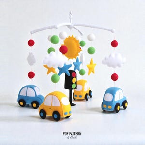 May include: A handmade baby mobile featuring felt cars, stars, clouds, and a sun. The mobile includes red, green, and white felt balls, and a traffic light. The cars are blue and yellow. The mobile is suspended from a white frame.