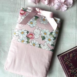 Peut inclure: Une housse de livre rose avec un motif floral composé de marguerites blanches, de roses roses et de lapins blancs. La housse est ornée d'un ruban de satin rose.