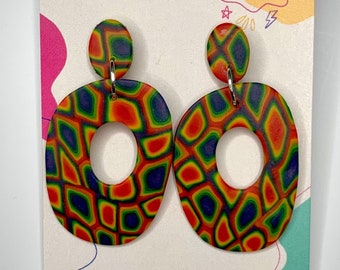 Puede incluir: Un par de pendientes ovalados, geométricos y coloridos con un agujero en el centro. Los pendientes están hechos de arcilla polimérica y tienen un patrón psicodélico de rojo, naranja, amarillo, verde y azul.