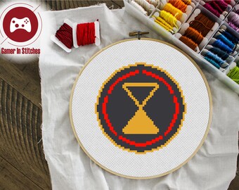 May include: A cross-stitch pattern of a black and gold hourglass with a red circle around it. The pattern is on white Aida cloth in a wooden embroidery hoop.