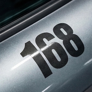 Könnte beinhalten: Nahaufnahme einer Autotür mit der Nummer "168" in einem schwarzen Kohlefaser-Muster. Die Zahlen sind groß und heben sich von einer hellgrauen Metalloberfläche ab. Die Autotür hat ein schlankes, modernes Design.