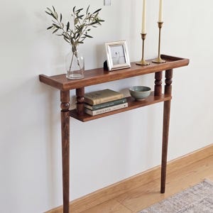 Può includere: Un tavolino consolle in legno marrone scuro con un ripiano inferiore. Il tavolo ha due candelabri in ottone con candele bianche, una piccola cornice, un vaso di vetro con verde e una piccola ciotola. Il tavolo è lungo circa 91 cm.