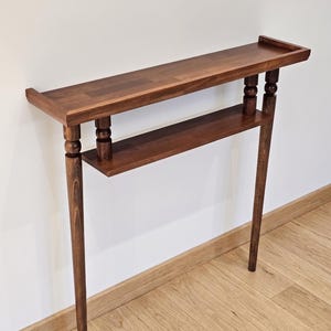 May include: A dark brown wooden console table with two shelves. The top shelf has a raised edge. The table has four legs, with a decorative element near the top of each leg. The table is against a white wall and wooden floor.