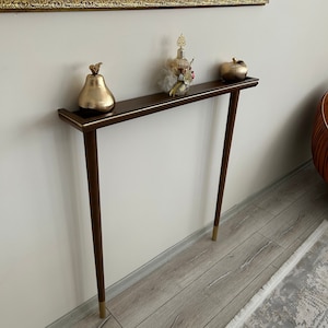 Puede incluir: Una mesa consola de color marr&oacute;n oscuro con detalles dorados y dos patas. La mesa tiene una pera dorada, una botella de perfume y una manzana dorada en la parte superior.