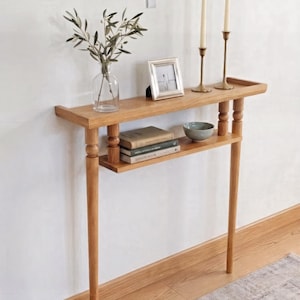 May include: A light brown wooden console table with a shelf. The table has two levels, with a shelf in the middle. On the top shelf are two brass candle holders with candles, a small picture frame, and a glass vase with greenery. The table is 122 cm long.