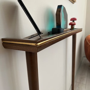 Puede incluir: Una mesa consola de madera marr&oacute;n oscuro con ribete dorado y dos patas. La mesa tiene una parte superior curva y est&aacute; decorada con una escultura de resina azul y marr&oacute;n, un soporte de tel&eacute;fono con un tel&eacute;fono negro y dos figuritas de setas rojas y blancas.