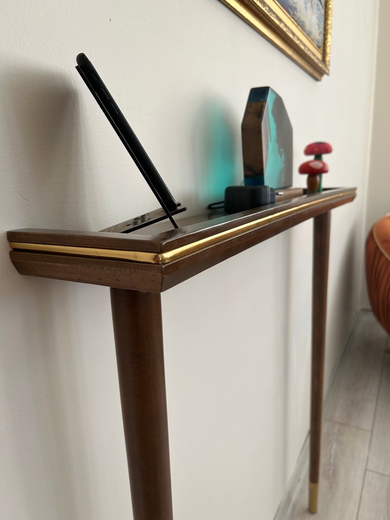 Puede incluir: Una mesa consola de madera marr&oacute;n oscuro con ribete dorado y dos patas. La mesa tiene una parte superior curva y est&aacute; decorada con una escultura de resina azul y marr&oacute;n, un soporte de tel&eacute;fono con un tel&eacute;fono negro y dos figuritas de setas rojas y blancas.