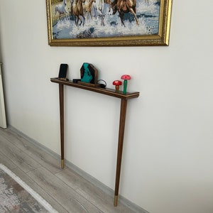 Puede incluir: Una mesa consola de madera con una tapa marr&oacute;n y patas con puntas doradas. La mesa est&aacute; decorada con una escultura de resina azul y verde, un tel&eacute;fono m&oacute;vil negro y tres figuritas de setas de madera.