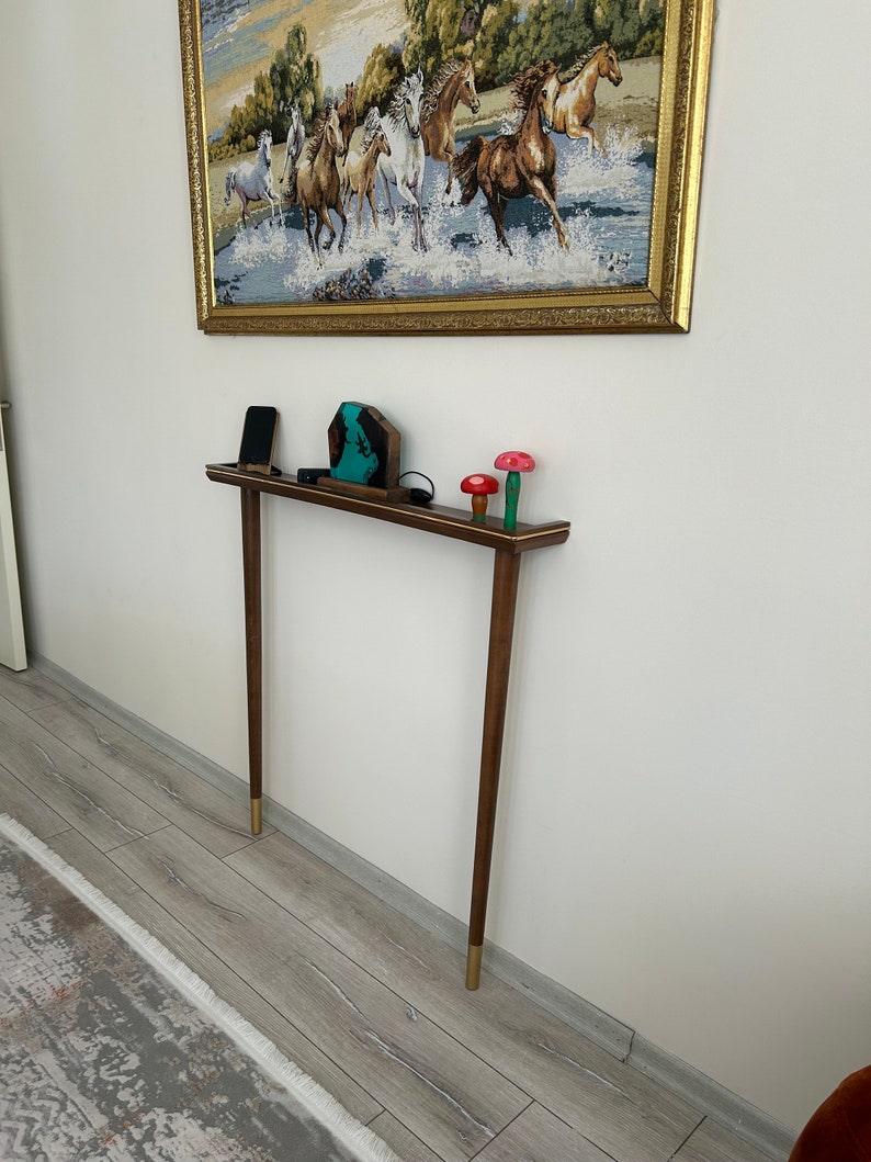 Puede incluir: Una mesa consola de madera con una tapa marr&oacute;n y patas con puntas doradas. La mesa est&aacute; decorada con una escultura de resina azul y verde, un tel&eacute;fono m&oacute;vil negro y tres figuritas de setas de madera.