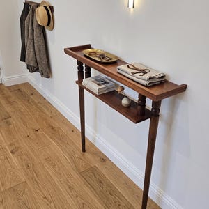 May include: A dark wood console table with two shelves. The top shelf holds a brass tray with keys and a folded linen cloth. The lower shelf displays a stack of books and a small vase with a flower. The table has long, slender legs.