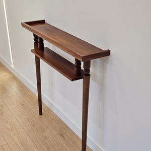 May include: A dark brown, two-tiered console table with a rectangular top and a lower shelf. The table features turned legs and decorative elements. The table is designed to be mounted against a wall.