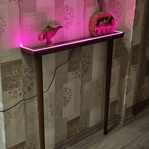 May include: A wooden console table with a pink LED light strip along the top edge. A gold-colored genie lamp and a decorative wooden vase with dried flowers sit on the table. The table has four slender legs and is against a patterned wallpaper.