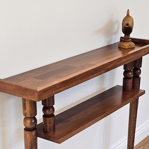 May include: A dark brown wooden console table with two shelves and turned legs. A decorative wooden bird sculpture sits on the top shelf. The table has a rectangular shape and is made of solid wood.
