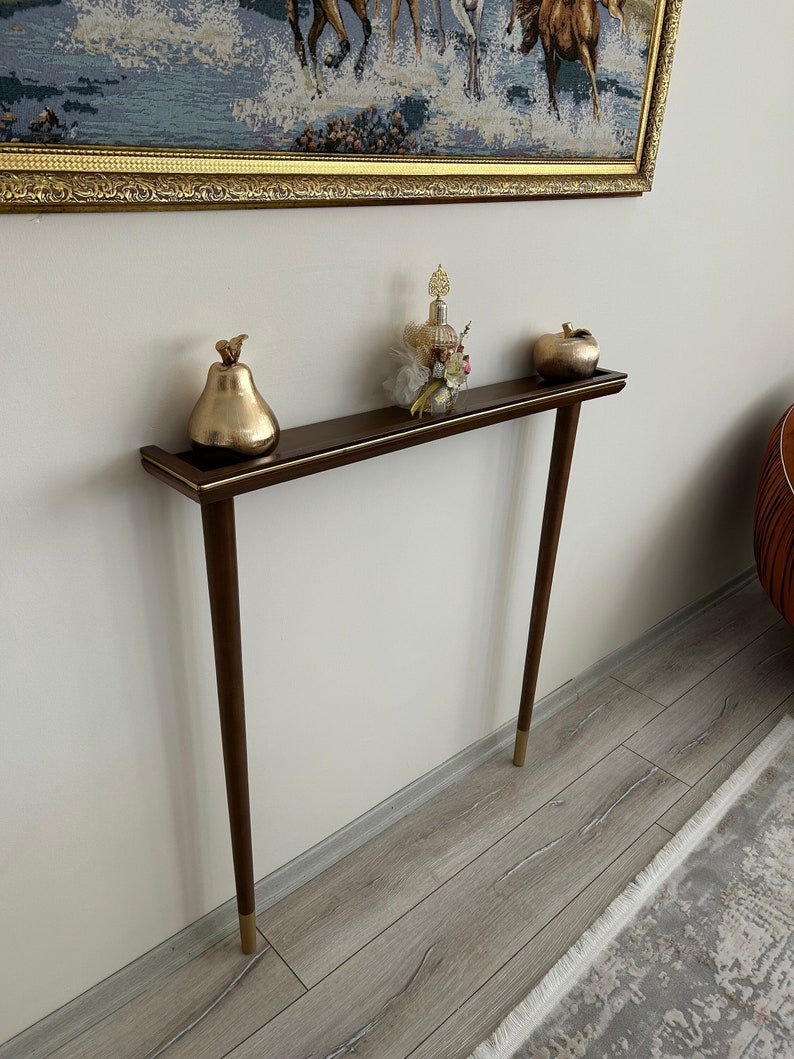 Puede incluir: Una mesa consola de color marr&oacute;n oscuro con detalles dorados y dos patas. La mesa tiene una pera dorada, una botella de perfume y una manzana dorada en la parte superior.