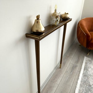 Puede incluir: Una consola de madera con una tapa marr&oacute;n y detalles dorados. La consola tiene dos patas y un estante. Hay art&iacute;culos decorativos en el estante, incluyendo una pera dorada, una botella de perfume y una manzana dorada.
