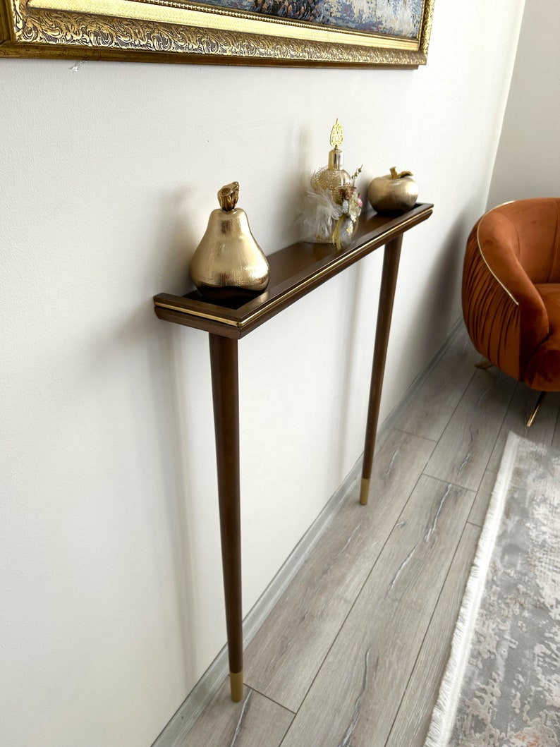 Puede incluir: Una consola de madera con una tapa marr&oacute;n y detalles dorados. La consola tiene dos patas y un estante. Hay art&iacute;culos decorativos en el estante, incluyendo una pera dorada, una botella de perfume y una manzana dorada.