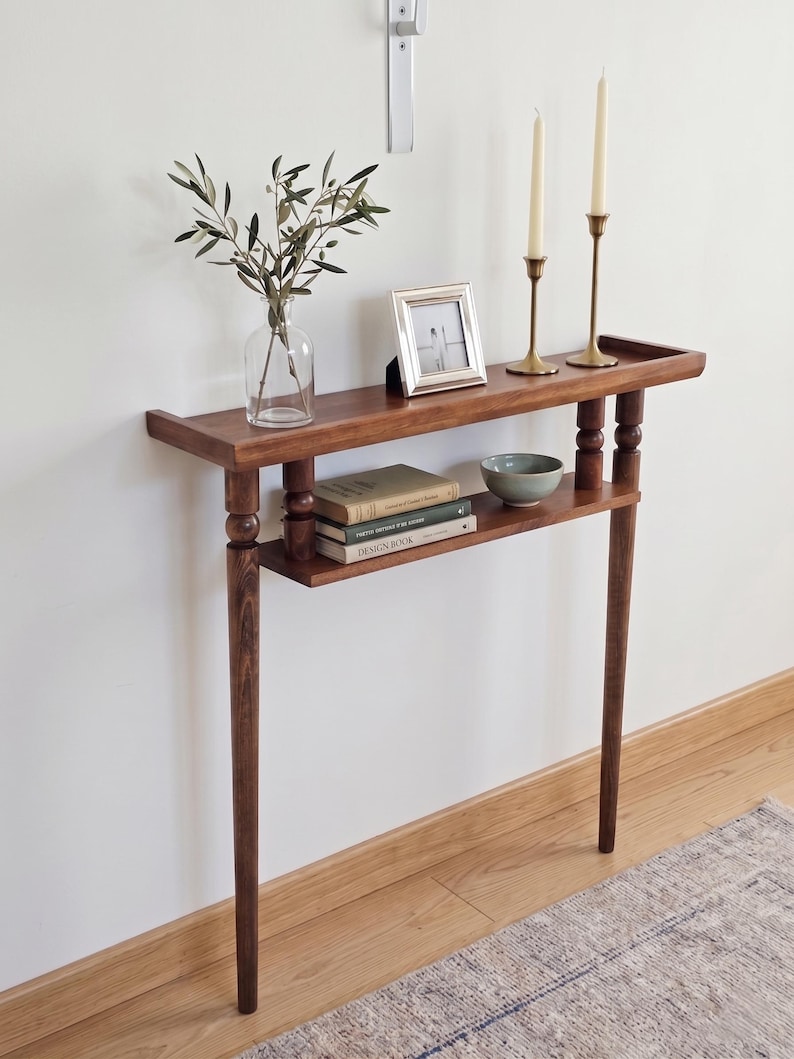 May include: A dark brown wooden console table with a lower shelf. The table has two brass candlesticks with white candles, a small picture frame, a glass vase with greenery, and a small bowl. The table is approximately 36 inches long.