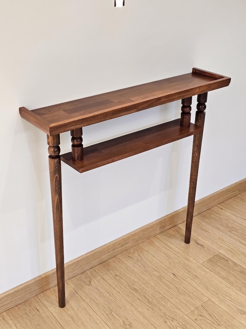 May include: A dark brown wooden console table with two shelves. The top shelf has a raised edge. The table has four legs, with a decorative element near the top of each leg. The table is against a white wall and wooden floor.