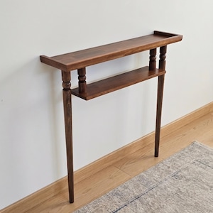 May include: A dark brown wooden console table with two shelves. The top shelf has a raised edge. The table has turned legs and a lower shelf. The table is against a white wall and sits on a wooden floor. A rug is partially visible.