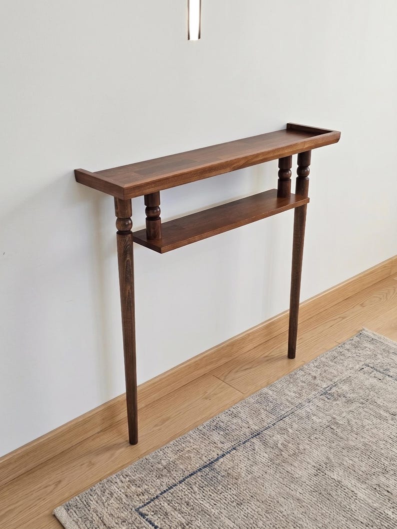 May include: A dark brown wooden console table with two shelves. The top shelf has a raised edge. The table has turned legs and a lower shelf. The table is against a white wall and sits on a wooden floor. A rug is partially visible.